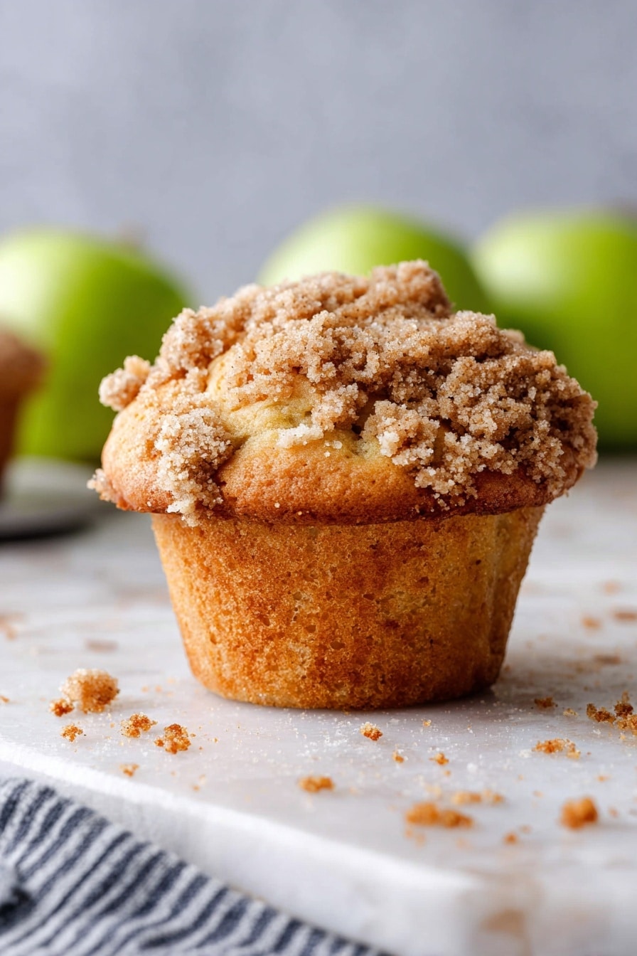 A dark metal muffin tray holds eleven freshly baked muffins, each topped with a thick layer of light brown crumbly streusel that is unevenly spread, giving a rough texture and scattered crumbs around the tray. The muffins themselves show a golden brown base with soft, creamy white swirls underneath the crumb topping. Around the tray, on a white marbled surface, a green apple sits near the top left corner, and a white and dark striped cloth lies folded on the right side. The scene is bright with soft natural light highlighting the textures of the muffins and crumbs. Photo taken with an iphone --ar 2:3 --v 7 - Apple Crumb Muffins, apple crumb muffins recipe, easy apple muffin ideas, cinnamon apple muffins, homemade apple muffins