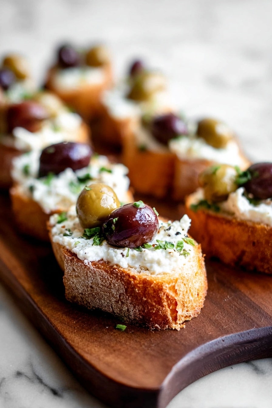 The image shows a small piece of toasted bread as the base layer with a golden-brown, crispy crust and soft, airy inside, topped thickly with a smooth white creamy spread. On top of the cream, there are whole olives in dark purple and green colors scattered along with small bits of fresh green herb. The toast with its toppings is placed on a dark wooden board that contrasts with the white marbled surface underneath. A bite is taken out of the toast, showing the airy texture inside. In the background, more of these toasts with the same toppings are blurred, giving a focus on the front piece. Photo taken with an iphone --ar 2:3 --v 7 - Whipped Feta Crostini Appetizer, easy feta crostini, quick party appetizers, flavorful crostini recipes, elegant snack ideas