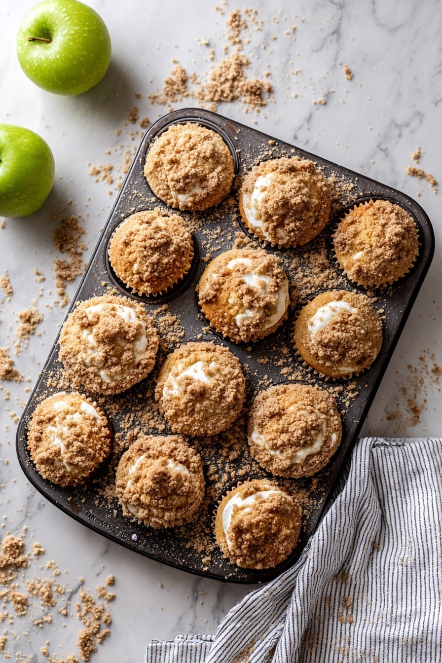The image shows eight crumb-topped muffins, arranged casually on white parchment paper over a white marbled surface. The muffins have a golden brown crumb layer on top, which is rough and textured, giving them a crunchy look. One muffin is broken in half, showing a soft, light, and fluffy yellow interior with crumb pieces around it. Two green apples, shiny and smooth, rest next to the muffins toward the left side of the image. Some crumbs are scattered around the muffins and apples, adding to the casual, homemade feel. A corner of a gray and white striped cloth is visible in the bottom left corner. Photo taken with an iphone --ar 2:3 --v 7 - Apple Crumb Muffins, apple crumb muffins recipe, easy apple muffin ideas, cinnamon apple muffins, homemade apple muffins