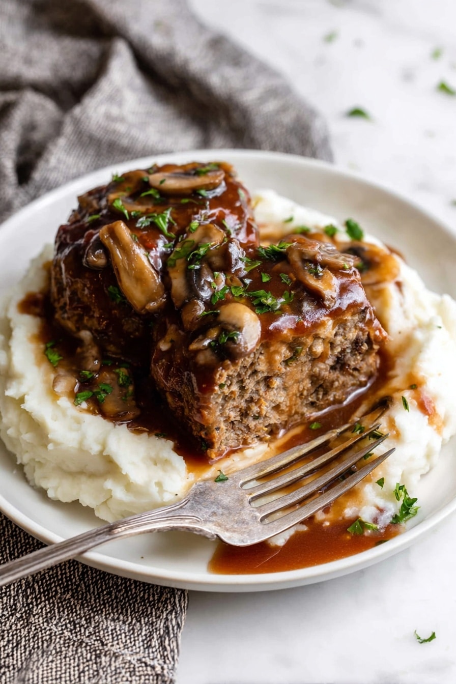 A white plate holds a layer of creamy white mashed potatoes spread evenly at the bottom. On top, there are two large portions of brown meatloaf, one of which is sliced open to show a dense, moist texture inside. The meatloaf is covered with a thick, rich brown sauce that has pieces of cooked mushrooms and bits of parsley scattered on top. A silver fork rests in the sliced meatloaf, pushing it slightly to the side, with a small amount of sauce dripping onto the mashed potatoes. The plate is placed on a white marbled surface with a greyish woven cloth nearby. Photo taken with an iphone --ar 2:3 --v 7 - Crock Pot Salisbury Steak with Mushroom Gravy, easy crock pot recipes, hearty beef dinner, comfort food recipes, slow cooker steak and gravy