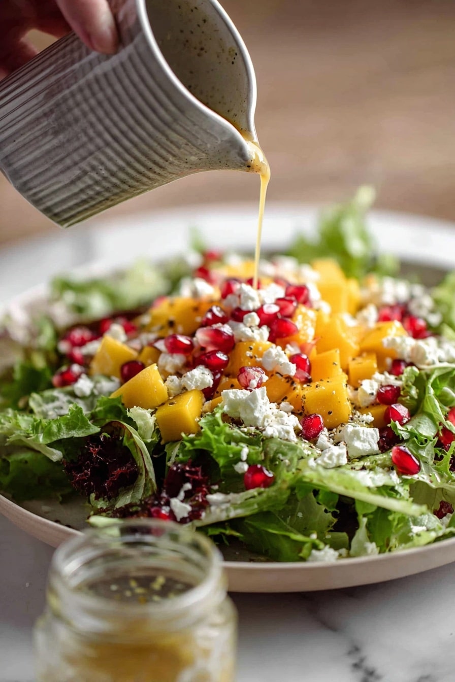 A white plate holds a fresh salad with three main layers: the bottom layer is mixed green lettuce leaves with curly edges, the middle layer has bright orange mango cubes, and the top layer is sprinkled with small white cheese crumbles and red pomegranate seeds. A woman's hand is pouring creamy light brown dressing from a gray ribbed pitcher onto the salad. A small glass jar with leftover dressing sits near the salad on a white marbled surface. Photo taken with an iphone --ar 2:3 --v 7 - Festive Pomegranate Walnut Salad, holiday salad recipes, easy holiday salads, colorful fruit and nut salads, healthy holiday side dishes