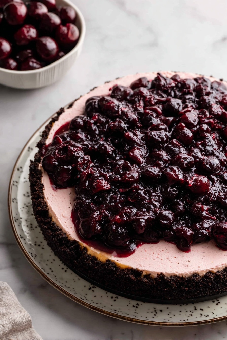 A three-layer round dessert sits on a white plate with a subtle pattern. The bottom layer is a rough dark chocolate crust with a crumbly texture, tightly packed and thick. The middle layer is a smooth, pale pink cream that fills the whole crust evenly. The top layer is a dark red cherry topping, thick and glossy, made of whole and crushed cherries, spread in an uneven, chunky way covering the cream completely. To the upper left corner, a white bowl is filled with whole dark red cherries. The scene is set on a white marbled surface. Photo taken with an iphone --ar 2:3 --v 7 - Cherry Cheesecake with Oreo Crust, cherry cheesecake with oreo crust, best cherry cheesecake dessert, easy cherry cheesecake recipe, homemade cherry cheesecake