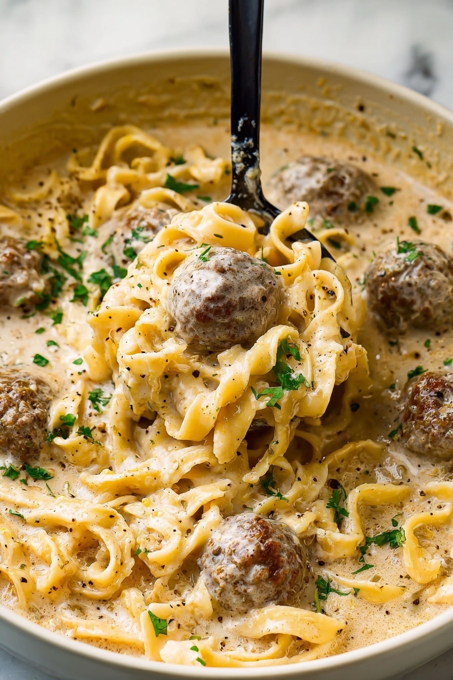 A close-up of a bowl filled with creamy pasta and meatballs in a rich beige sauce. The bowl is white with a dark rim, sitting on a white marbled surface. Inside, there are two main layers: the base layer of twisted pale yellow pasta with smooth texture, and on top, several round, browned meatballs covered in smooth, thick light cream sauce flecked with black pepper and small green herbs. A gold spoon holds one meatball, partially submerged in sauce. Photo taken with an iphone --ar 2:3 --v 7 - Swedish Meatball Soup, easy Swedish meatball soup, comforting Swedish soup, cozy meatball soup recipe, homemade Swedish meatball soup