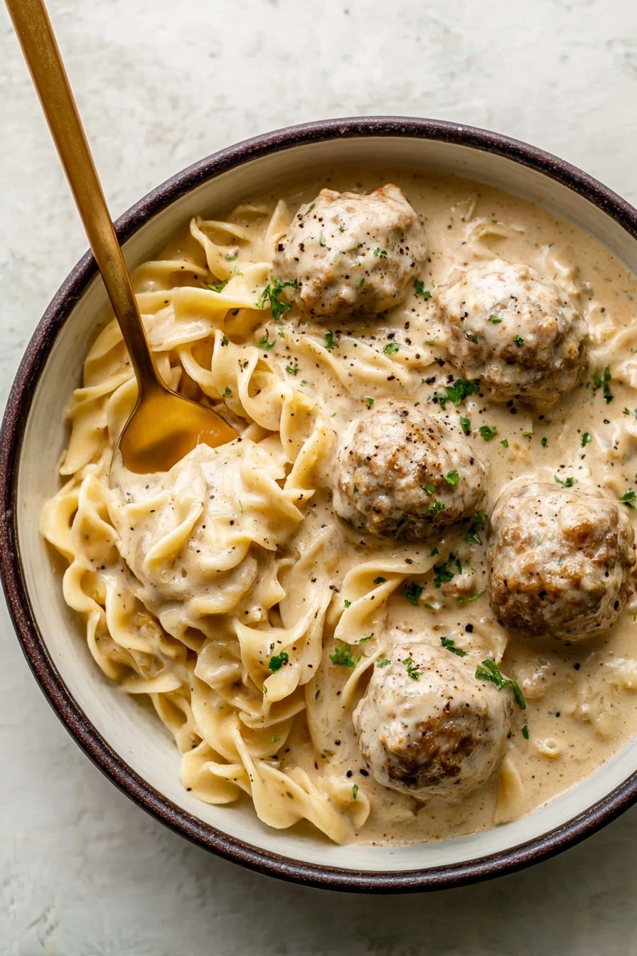 There is a close-up of a creamy noodle dish with meatballs in a white bowl. The dish has thick, pale yellow noodles that twist and curl, mixed in with round, browned meatballs. The creamy sauce is light beige with specks of black pepper and green herbs scattered over the top. A black ladle is lifting some noodles and a meatball, showing the thick sauce coating them. The surface beneath the bowl is a white marbled texture. photo taken with an iphone --ar 2:3 --v 7 - Swedish Meatball Soup, easy Swedish meatball soup, comforting Swedish soup, cozy meatball soup recipe, homemade Swedish meatball soup