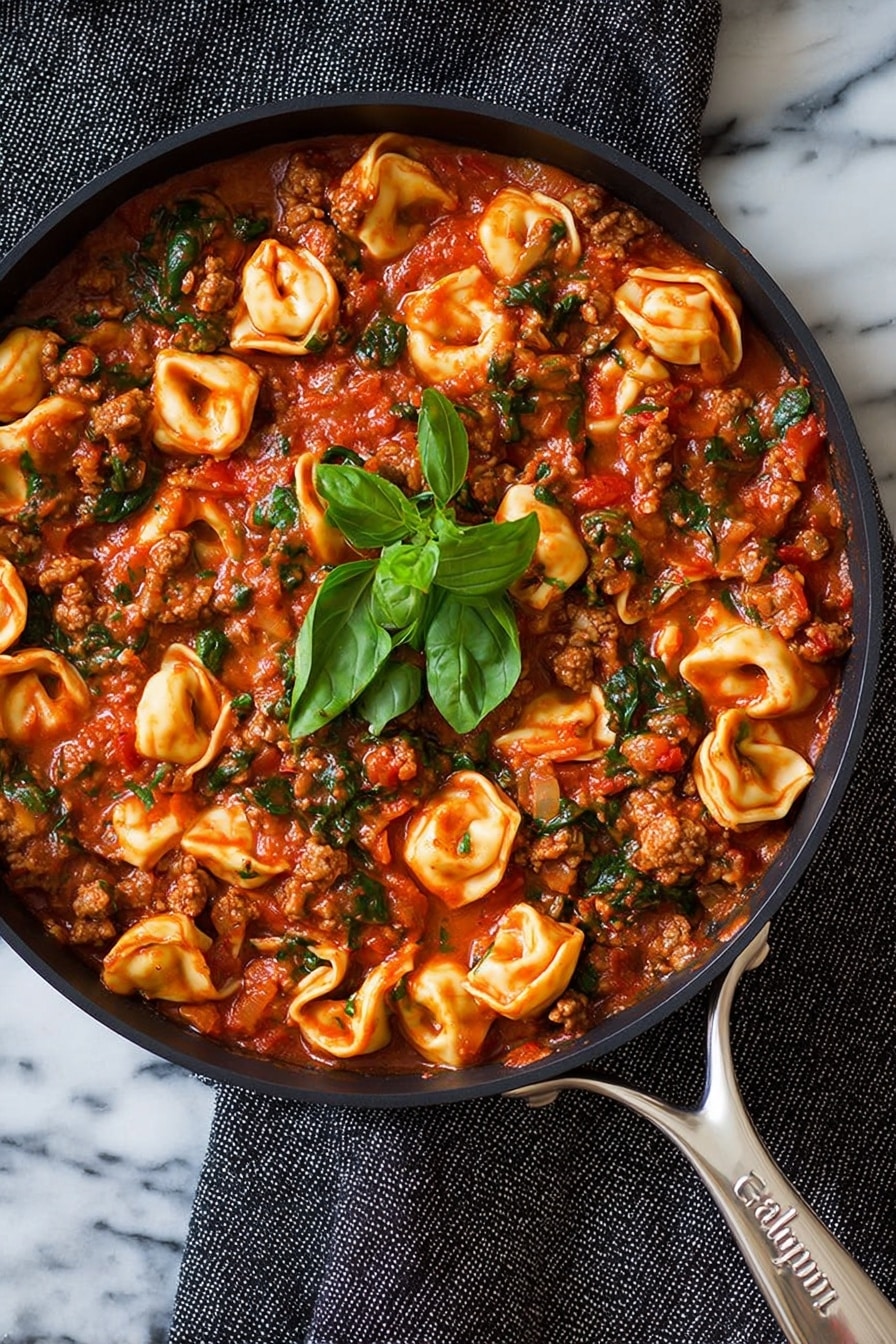 The image shows a close-up of a black skillet filled with tortellini pasta cooked in a rich red tomato sauce mixed with small pieces of cooked meat and chopped green spinach, all evenly spread throughout. On top in the center, there is a small bunch of fresh green basil leaves. The skillet rests on a white marbled surface with a dark gray textured cloth nearby. The shiny silver handle of the skillet is visible in the bottom right corner with the brand name Calphalon engraved on it. Photo taken with an iphone --ar 2:3 --v 7 - One-Pan Turkey Sausage and Spinach Tortellini, Turkey Sausage Tortellini Skillet, Easy Turkey Sausage Pasta, Cheeseburger Spinach Tortellini, Quick Weeknight Pasta