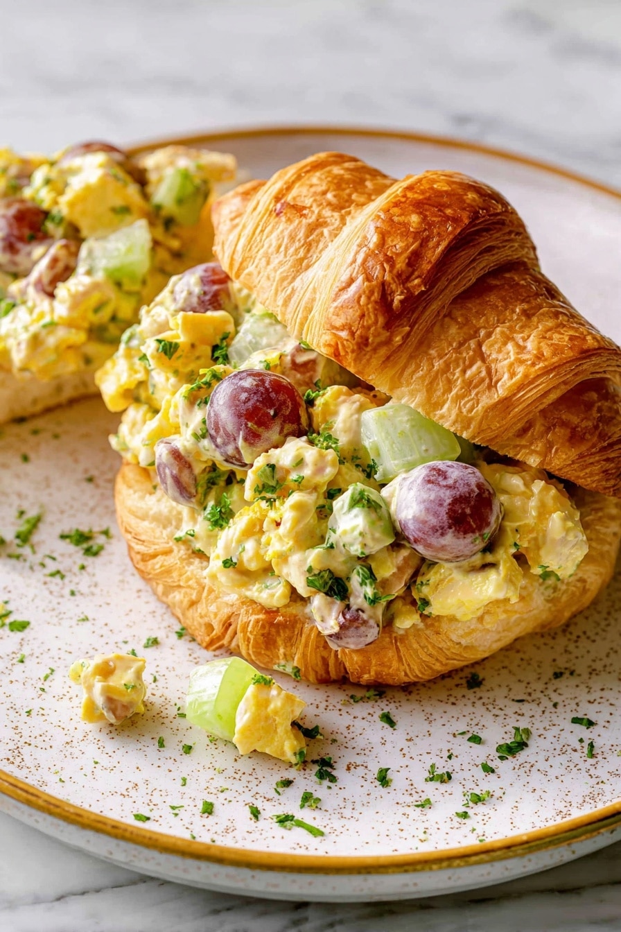 A flaky, golden-brown croissant is sliced in half and filled with a colorful chicken salad. The salad has chunks of light yellow chicken mixed with small pieces of pale green celery, and whole small round purple grapes, all coated in a creamy yellow dressing. Some chopped green herbs are sprinkled on top and on the white speckled plate with a gold rim beneath it. The background is a white marbled texture photo taken with an iphone --ar 2:3 --v 7 - Creamy Chicken Curry Salad, chicken curry salad, chicken salad with curry mayo, easy chicken salad recipes, flavorful chicken salad