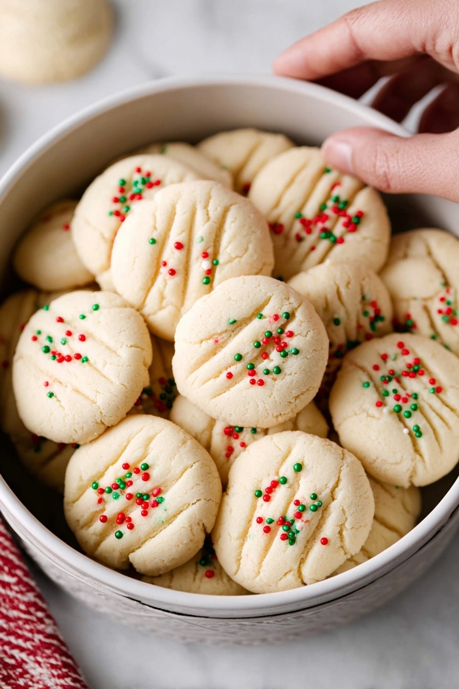 The image shows a stack of four soft, pale beige cookies standing on a white marbled surface, with the top cookie broken in half to reveal a crumbly, dense inside. Each cookie is round and smooth, topped with small, red and green sprinkles that add a festive touch. The stack is very close to the camera, with the background softly blurred and colors muted. Photo taken with an iphone --ar 2:3 --v 7 - Whipped Shortbread Cookies, melt-in-your-mouth shortbread, buttery cookies recipe, fluffy shortbread dough, holiday cookie recipes