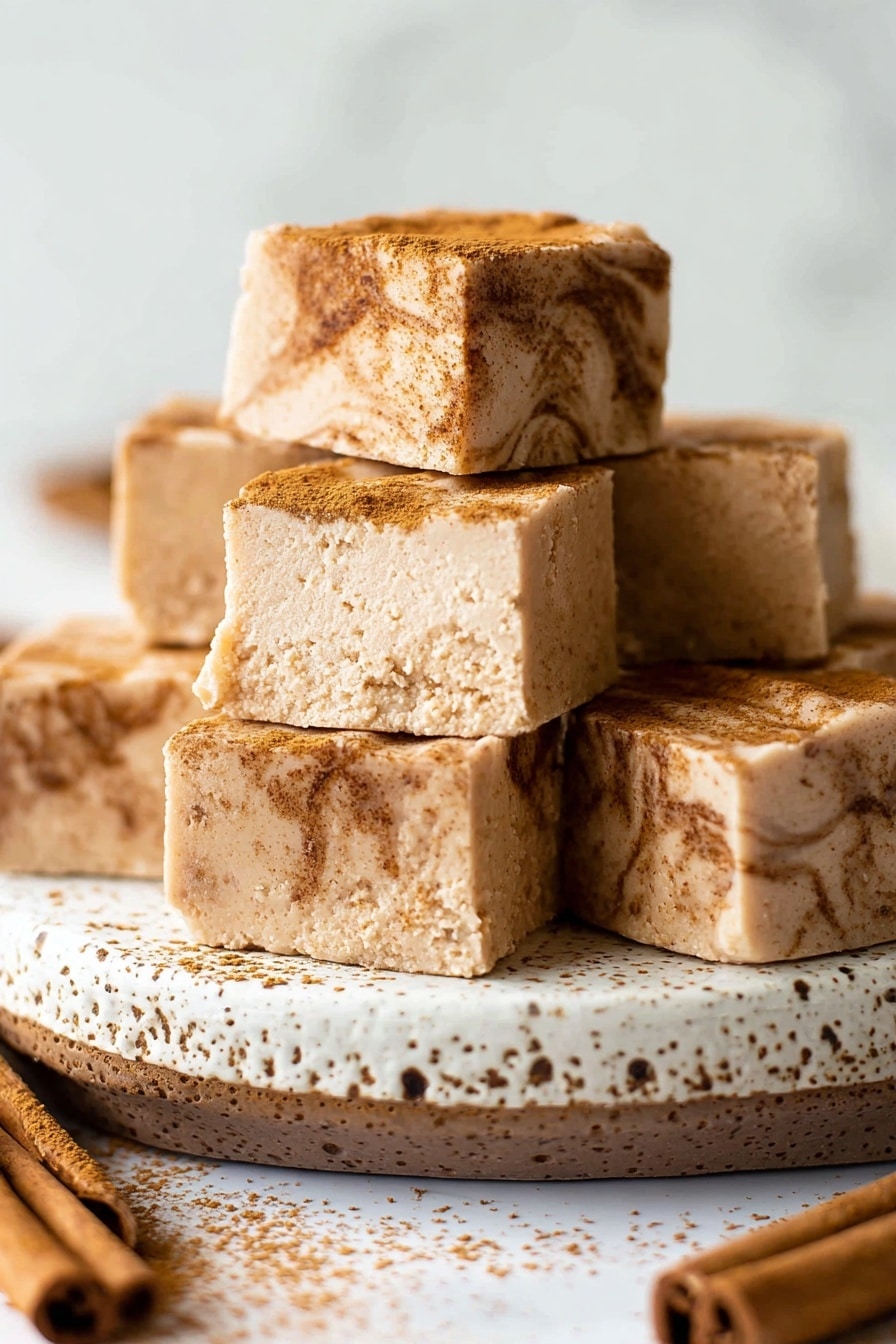 The image shows a stack of soft, square-shaped dessert pieces arranged on a white plate with a slightly speckled pattern. There are two layers visible in the dessert: the bottom layer is light brown and smooth, while the top layer is darker brown with a crumbly texture that looks like crushed nuts or cinnamon. The stack consists of six squares, with three squares at the bottom, two in the middle, and one on top. The background and surface are a white marbled texture, giving the image a clean and bright look. photo taken with an iphone --ar 2:3 --v 7 - Cinnamon Sugar White Chocolate Fudge, white chocolate fudge, cinnamon fudge recipe, easy holiday fudge, homemade fudge dessert