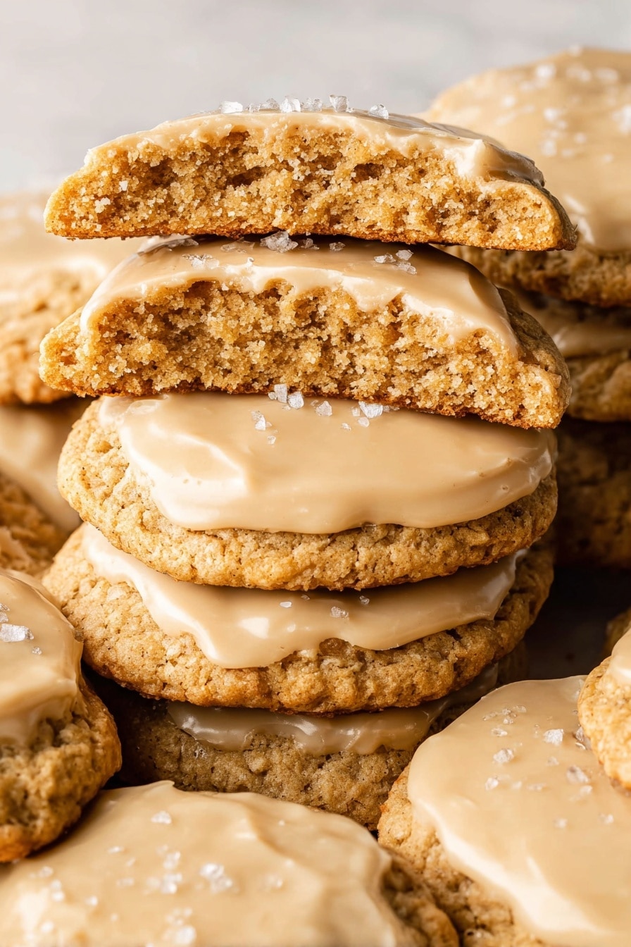 A close-up of a stack of soft, golden-brown cookies with a slightly rough, crumbly texture, each sandwiching a thick layer of smooth, creamy light caramel-colored frosting. The top cookie on the front stack is bitten, showing its dense inner texture and frosting layer inside. The frosting on the top cookies is spread thickly and has small white flakes sprinkled on top, likely sea salt. The cookies rest on a white marbled surface, with the stacks leaning slightly against each other, showing multiple layers of cookies and frosting. photo taken with an iphone --ar 2:3 --v 7 - Salted Caramel Cookies, salted caramel cookies recipe, chewy caramel cookies, buttery caramel cookies, salted caramel cookie ideas