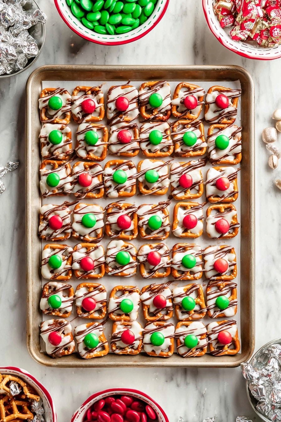 A baking tray holds 72 small square pretzels arranged in a grid. Each pretzel has a white, melted candy layer on top, with thin dark brown stripes drizzled over it. On top of the candy layer, a single large red or green candy is placed in the center of each pretzel, alternating colors in rows. The tray sits on a white marbled surface. Around the tray, there are three bowls: one is white with a green rim holding red and green candies, and the other two are white with red rims, one with extra square pretzels and the other with some wrapped silver candies. photo taken with an iphone --ar 2:3 --v 7 - Chocolate Pretzel Hugs with M&Ms, easy holiday treats, no-bake chocolate snacks, salty sweet pretzel bites, festive chocolate M&Ms desserts