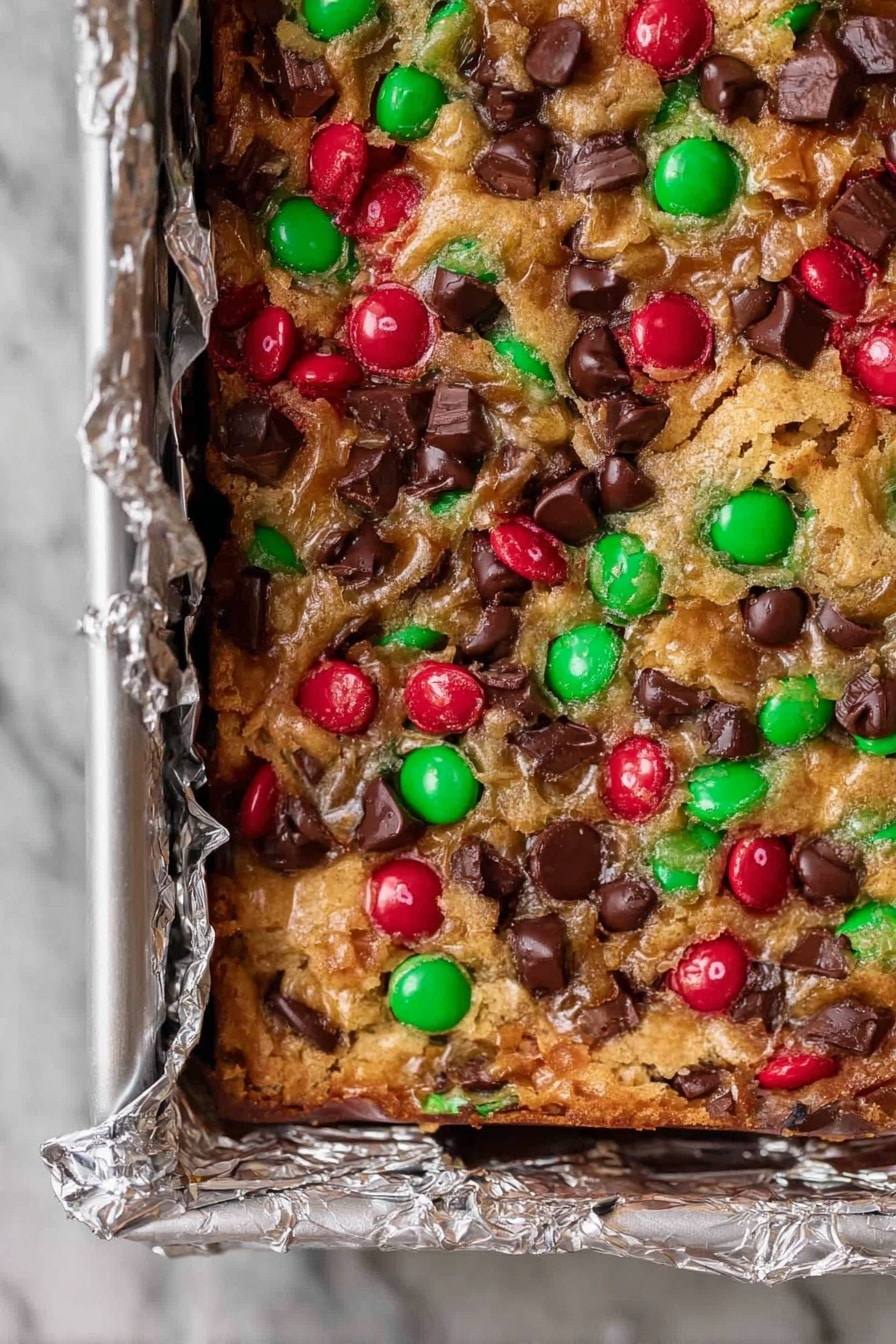 A close-up view of a square tray lined with foil filled with a baked dessert bar. The top layer shows a mix of red and green candy-coated chocolates and dark brown chocolate chips scattered unevenly on a golden-brown, slightly melted and crispy textured base. The surface of the bar looks bubbly and crunchy, with the bright candy colors breaking up the rich chocolate tones, filling the frame and showing a dense, festive-looking treat. The tray rests on a white marbled texture. Photo taken with an iphone --ar 2:3 --v 7 - Christmas Magic Cookie Bars, holiday cookie bars, festive dessert recipes, easy Christmas treats, chocolate coconut M&M's bars