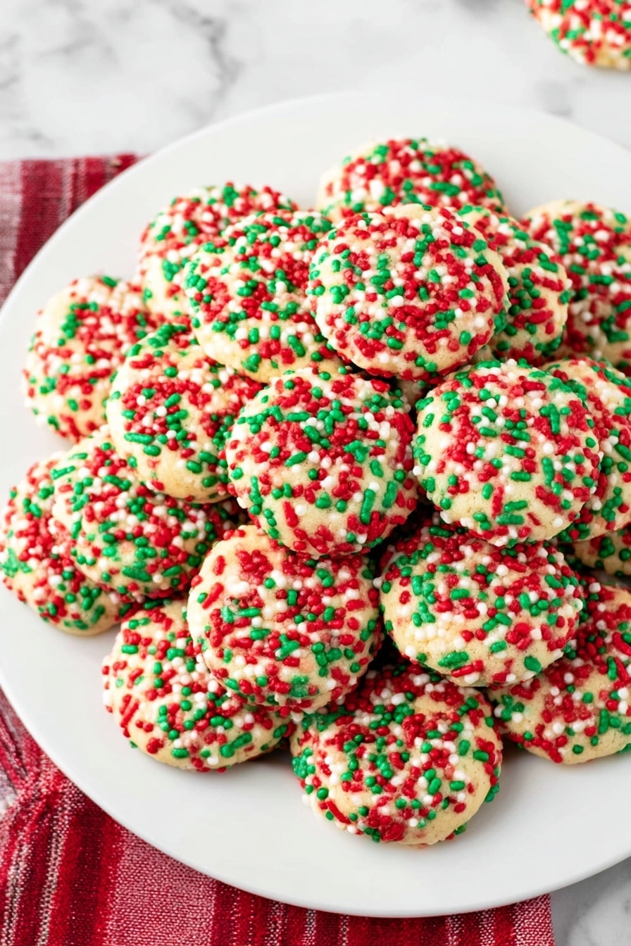 The image shows a white plate on a white marbled surface holding a stack of four round cookies covered fully in red, green, and white sprinkles. The cookies are pale yellow with a slightly bumpy texture beneath the colorful, small cylindrical sprinkles that stick out evenly in all directions. Some sprinkles are scattered around the plate, adding a festive look. In the background, there is a blurred white cup and red checkered fabric. The lighting is bright and natural, highlighting the vibrant colors of the sprinkles. photo taken with an iphone --ar 2:3 --v 7 - Christmas Sprinkle Pudding Cookies, festive holiday cookies, chewy vanilla pudding cookies, colorful Christmas cookie recipes, easy holiday cookie ideas
