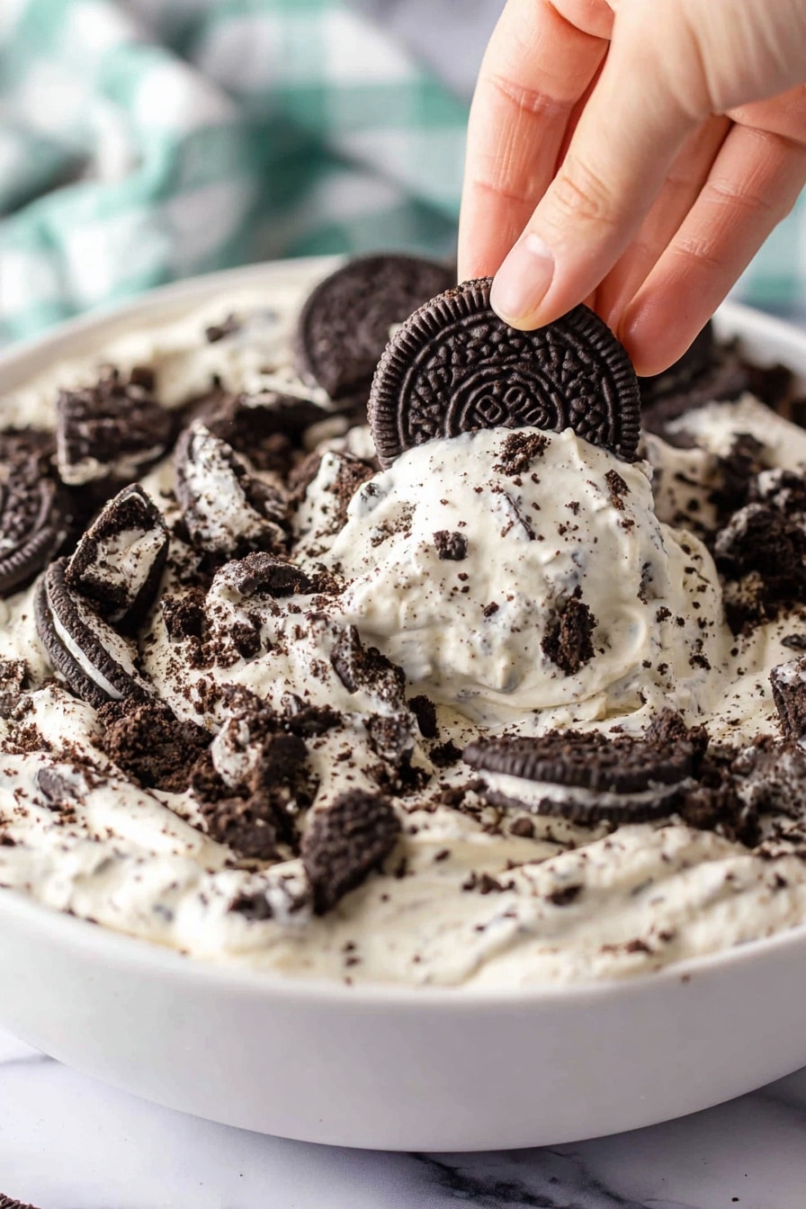A close-up view shows a white bowl filled with a creamy white mixture layered generously with dark chocolate sandwich cookie pieces. The creamy layer has a smooth, thick texture with crushed cookie bits mixed in, giving it a speckled look. On top, larger broken cookie pieces are spread unevenly, some slightly dipped into the creamy layer. A woman's hand is holding a whole dark chocolate sandwich cookie partially dipped in the creamy mixture, lifting it from the bowl. The background displays a white marbled surface and a blurred checkered cloth in soft colors. Photo taken with an iphone --ar 2:3 --v 7 - Oreo Fluff Dessert, no-bake Oreo dessert, quick Oreo fluff, creamy chocolatey dessert, crowd-pleasing Oreo treat
