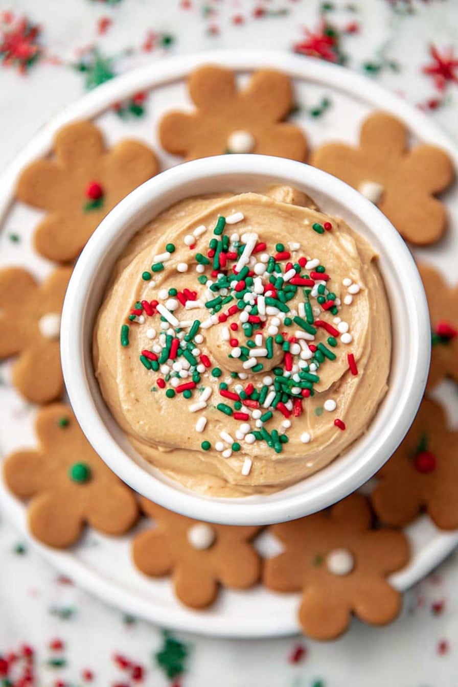 A white bowl filled with smooth, light brown dip topped with colorful Christmas-themed sprinkles including red, green, and white small balls, rods, and tree shapes. A large, round, golden-brown cookie is placed leaning on the top edge of the bowl. The bowl is set on a white marbled surface. Photo taken with an iphone --ar 2:3 --v 7 - Sweet Gingerbread Cheesecake Dip, festive holiday dip, easy gingerbread dip, creamy holiday appetizer, gingerbread flavored dip