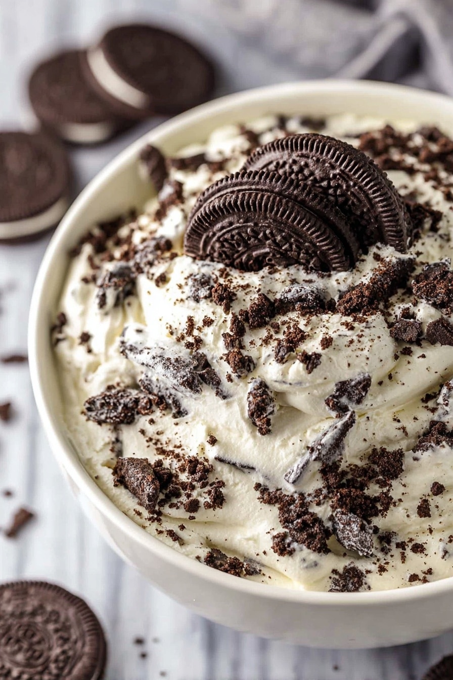 A close-up view of a white bowl filled with a creamy dessert covered in crushed chocolate sandwich cookies mixed throughout. The top layer is thick and white with creamy texture, generously sprinkled with small and large cookie pieces in dark brown and black shades. One whole sandwich cookie with visible cream center rests on top near the center, drawing focus. The bowl sits on a white marbled surface, and more whole cookies are blurred in the background. Photo taken with an iphone --ar 2:3 --v 7 - Oreo Fluff Dessert, no-bake Oreo dessert, quick Oreo fluff, creamy chocolatey dessert, crowd-pleasing Oreo treat