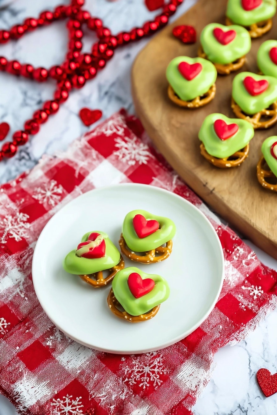 The image shows small snacks arranged in a diagonal line on a light wooden board on a white marbled surface. Each snack has two layers: the bottom half is green dipped coating with a smooth texture, and the top half is a light brown twisted pretzel shape with a rough texture. At the center of the pretzel is a whole almond. A small red heart-shaped decoration is placed on the green layer of each snack, adding a bright contrast. Around the board, there are scattered red heart shapes and a string of red beads on a red and white cloth. Photo taken with an iphone --ar 2:3 --v 7 - Grinch Pretzel Bites Caramel and Candy Melts, festive holiday treats, easy holiday snacks, green pretzel bites with caramel, Christmas party desserts