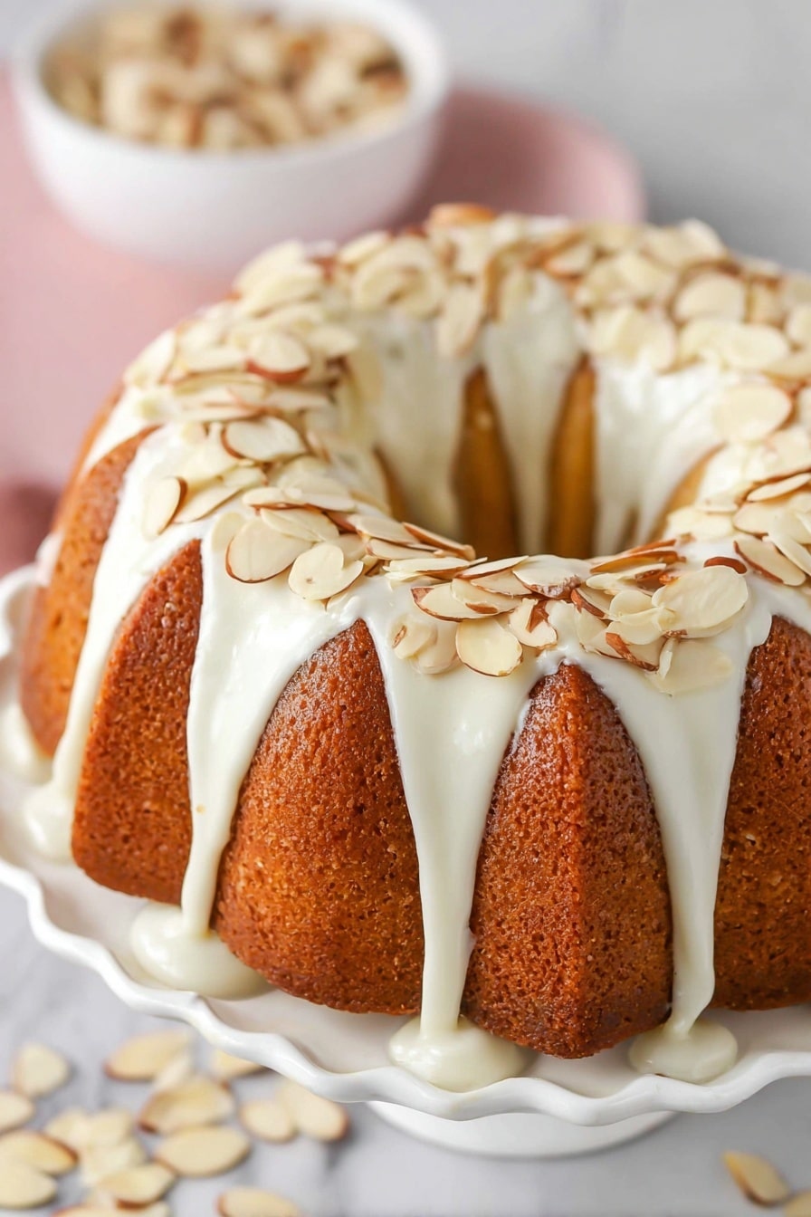 A round bundt cake with a moist, golden yellow inside and a light brown crust sits on a white cake stand. The cake has a thick layer of white icing that drips unevenly down the sides, topped with thin, light brown almond slices scattered on top and around the base. The cake stand has a glossy finish and a wide base, while the surface beneath shows a white marbled texture. A few crumbs and almond slices lie around the cake base. photo taken with an iphone --ar 2:3 --v 7 - Amaretto Almond Bundt Cake, almond bundt cake recipe, Amaretto cake, almond dessert, almond liquor cake