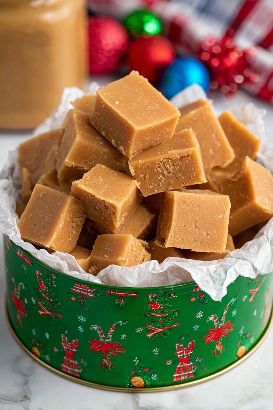 The image shows a round green tin with Christmas patterns, filled with many small square pieces of light brown fudge piled up inside. The fudge has a smooth texture with slight crumbly edges and is sitting on crumpled white parchment paper inside the tin. The background features a white marbled surface with blurry colorful Christmas ornaments and a peanut butter jar visible behind the tin. photo taken with an iphone --ar 2:3 --v 7 - Easy 2-Ingredient Peanut Butter Fudge, simple peanut butter fudge, quick peanut butter fudge, creamy peanut butter fudge, no-bake peanut butter fudge