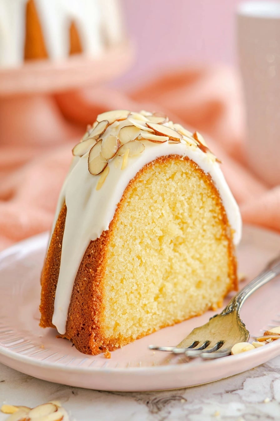 A slice of bundt cake stands on a white plate with soft pink tint, showing one thick layer of light yellow moist cake with a golden brown crust. The top of the slice is covered with a smooth, white icing layer, which holds sliced almonds that add texture and color contrast with their light cream and brown edges. A silver fork lies beside the cake slice on the plate. The surface beneath the plate has a white marbled texture with a soft peach cloth and blurred background in pastel tones. photo taken with an iphone --ar 2:3 --v 7 - Amaretto Almond Bundt Cake, almond bundt cake recipe, Amaretto cake, almond dessert, almond liquor cake