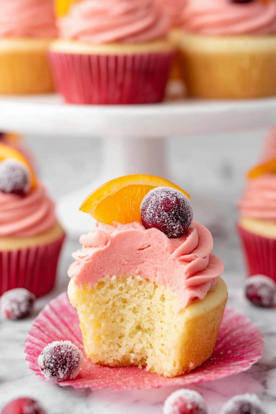 A group of light yellow cupcakes with three thick, smooth layers of bright pink frosting swirled in a spiral on top. Each cupcake is decorated with a thin orange slice, two red cranberries (one covered in sugar), and a light dusting of powdered sugar. The cupcakes are placed on a white cake stand and on a white marbled surface, with a red napkin under one cupcake in front. Scattered sugared cranberries are also on the surface around the cupcakes. The background has some blurred elements including a red cloth and glasses. photo taken with an iphone --ar 2:3 --v 7 - Cranberry Champagne Cupcakes, festive champagne cupcakes, holiday cranberry cupcakes, elegant cranberry dessert, sparkling cupcake recipes