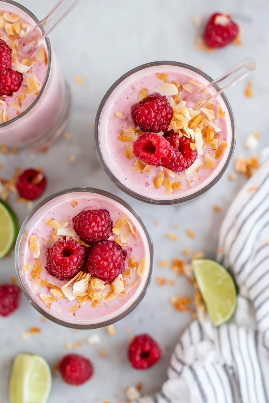 Three clear glass cups filled with a light pink smoothie are shown from above, each topped with bright red raspberries and small pieces of golden toasted flakes scattered on top. Each cup has a clear straw placed on the right side. The cups rest on a white marbled surface, with lime wedges and scattered toasted flakes visible around them. A white cloth with thin black stripes is seen on the right side of the image. photo taken with an iphone --ar 2:3 --v 7 - Mango Raspberry Smoothie with Chia Seeds, healthy mango raspberry smoothie, easy fruit smoothie recipes, nutritious chia seed smoothies, vibrant berry mango drink