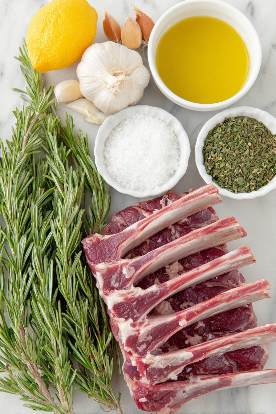 Flat lay of fresh rosemary sprigs with vibrant green needle-like leaves, four whole uncracked brown garlic cloves, a small white ceramic bowl filled with coarse kosher salt crystals, a small white ceramic bowl containing freshly ground black pepper, a whole medium lemon next to a heap of bright yellow lemon zest shavings, a small white ceramic bowl of freshly squeezed lemon juice, a small white ceramic bowl of golden extra virgin olive oil, two frenched racks of lamb with exposed clean white bones and rich red meat, all arranged symmetrically on a clean white marble surface, soft natural light, photo taken with an iPhone, professional food photography style, fresh ingredients, white ceramic bowls, no bottles, no duplicates, no utensils, no packaging --ar 2:3 --v 7 --p m7354615311229779997 - Garlic Rosemary Rack of Lamb, garlic rosemary lamb, roast lamb with garlic and rosemary, easy lamb recipe, flavorful rack of lamb